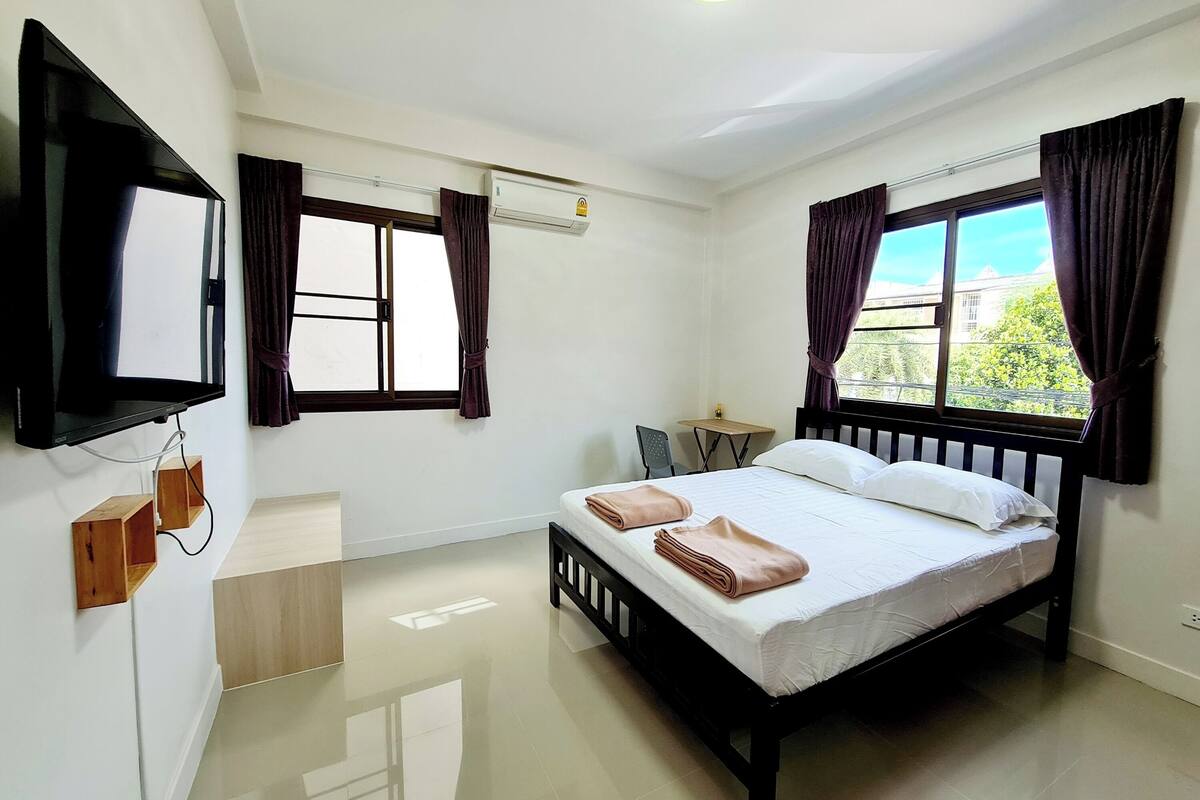 A clean, modern hotel room with a bed, a flat-screen TV mounted on the wall, a small table, and two windows with curtains.