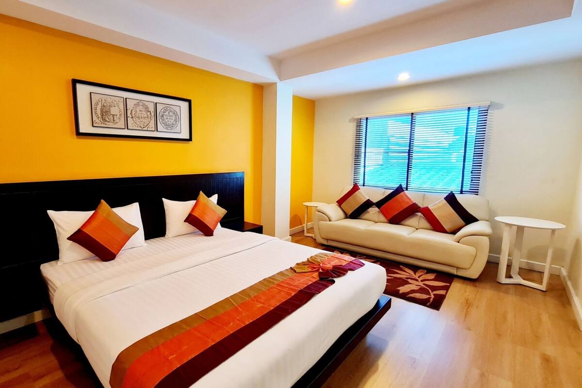 A hotel room with a large bed, a sofa, a small table, and a window with blinds.
