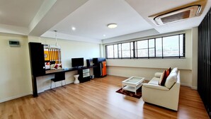 A modern living room with a sofa, coffee table, and a TV. There is a window with blinds, a ceiling fan, and a small kitchen area with a refrigerator. - TP House @town (Phuket)