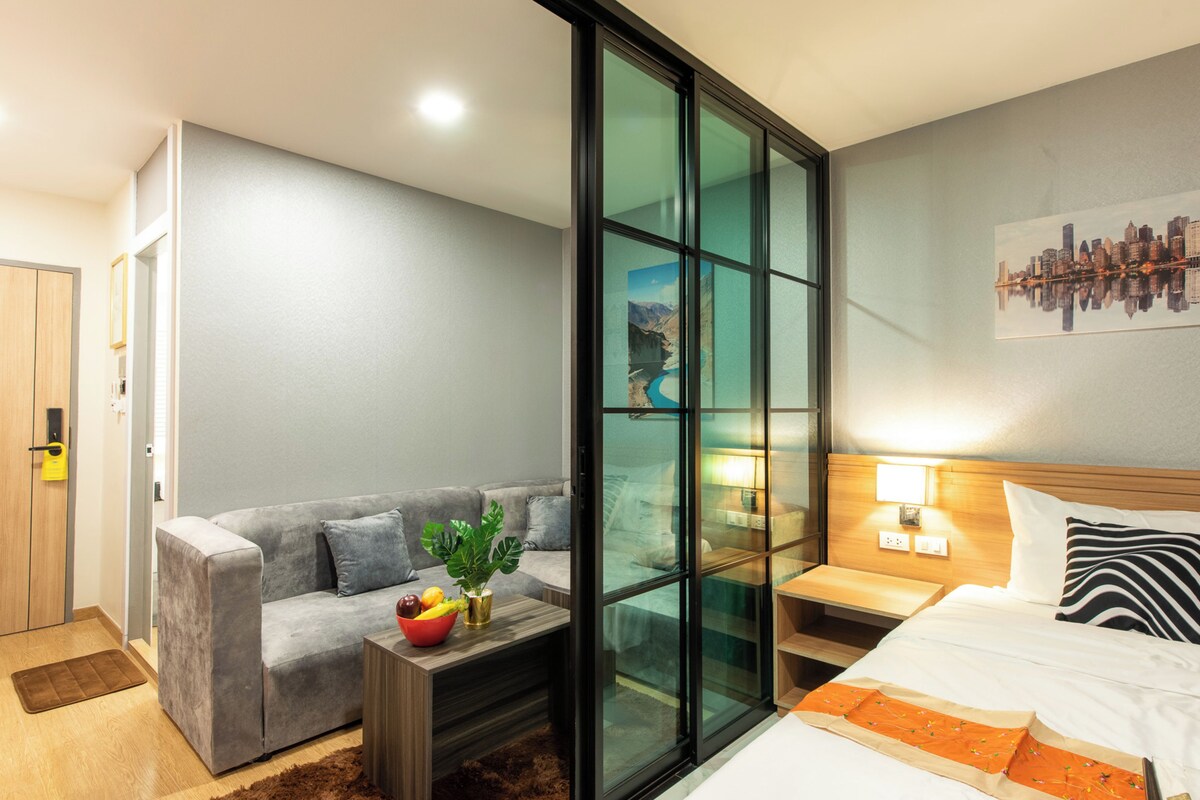 A modern hotel room with a grey sofa, a small coffee table with fruits, a bed with a striped pillow, and a cityscape view through a glass wall.