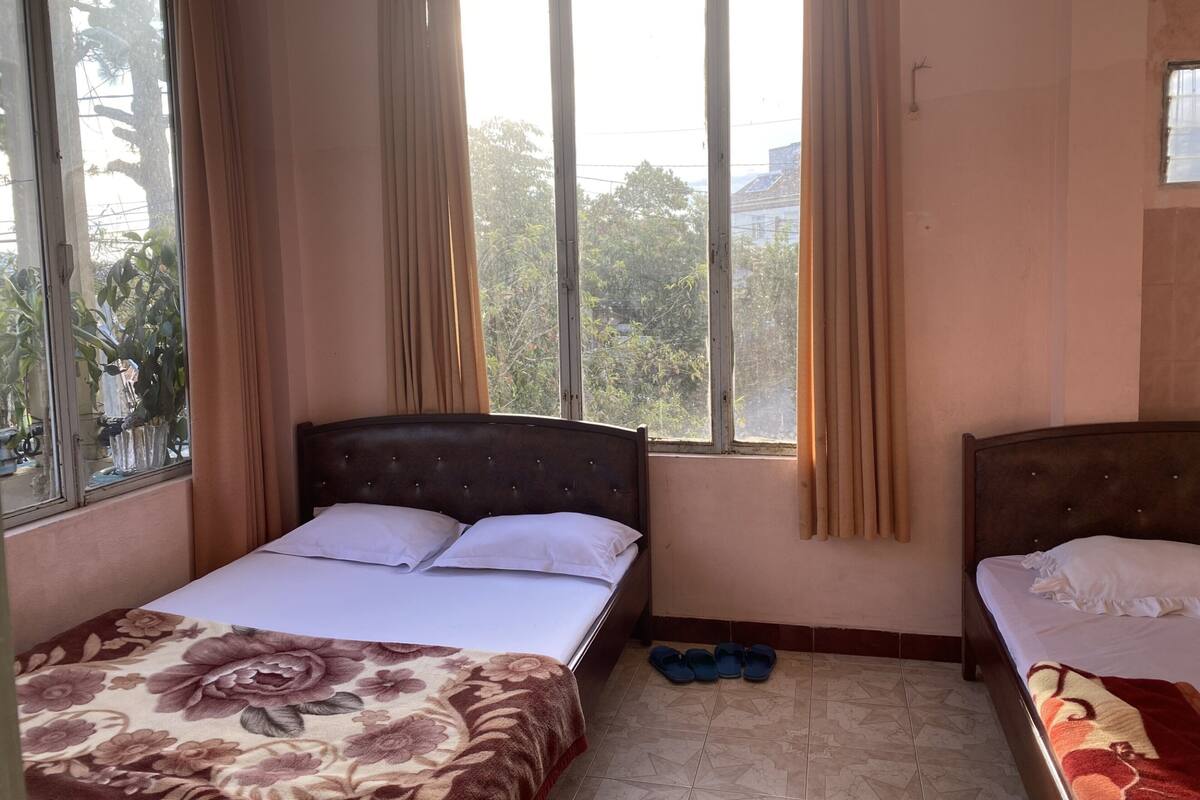 A room with two beds, a window with curtains, a tiled floor, and a pair of slippers on the floor.