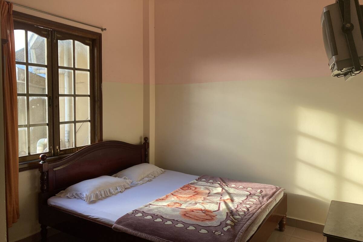 A single bed with a wooden headboard, a window with curtains, and a wall-mounted TV.