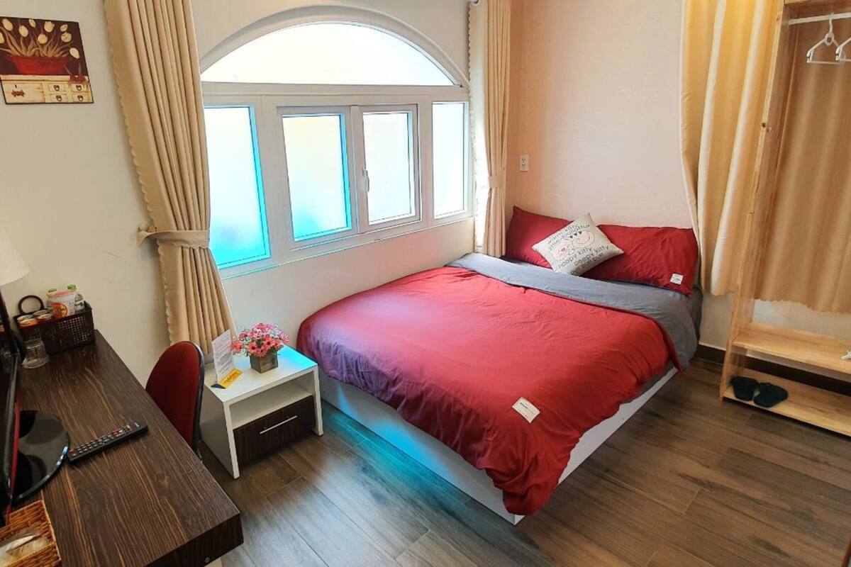 A hotel room with a bed, a desk with a TV, a small table with a vase of flowers, and a window with curtains.
