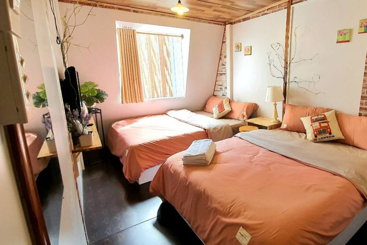 A cozy room with two beds, a window with curtains, a brick wall, and a small shelf with items.