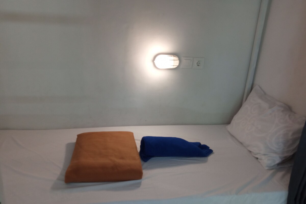 A single bed with a white mattress and two pillows, one brown and one blue, in a room with a light fixture on the wall.