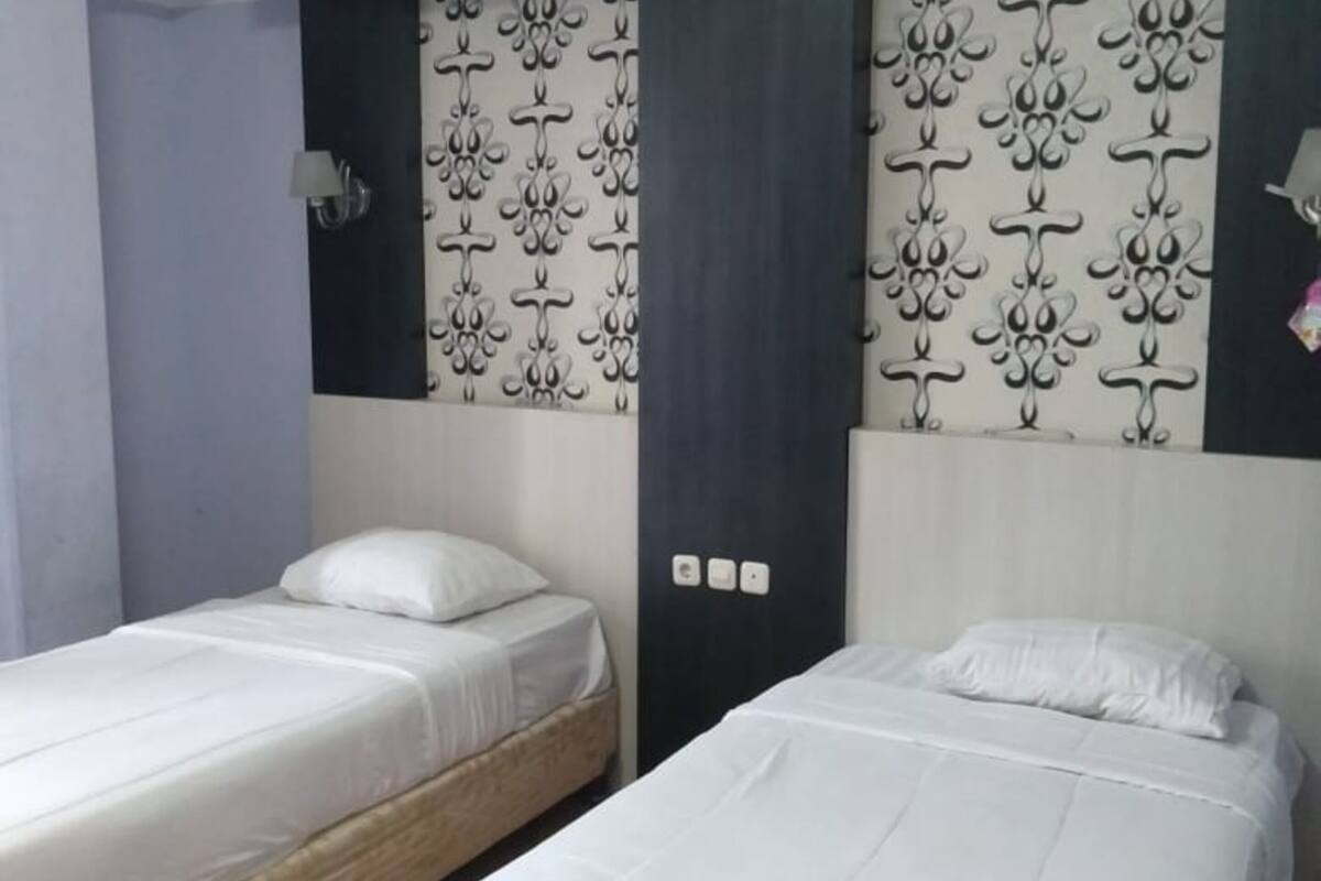 Ein Hotelzimmer mit zwei Einzelbetten, einem dunklen Kopfteil mit Verziierungsmuster und einer schlichten Wand mit Lichtschalter.