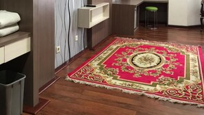 A room with a wooden floor, a red patterned rug, a TV mounted on the wall, and a small stool. - Grand Balqis Akasia Hotel (Surabaya)