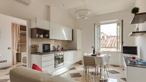 Una cocina moderna con armarios blancos, un comedor con mesa y sillas, y vista a un tejado con baldosas a través de una ventana.
