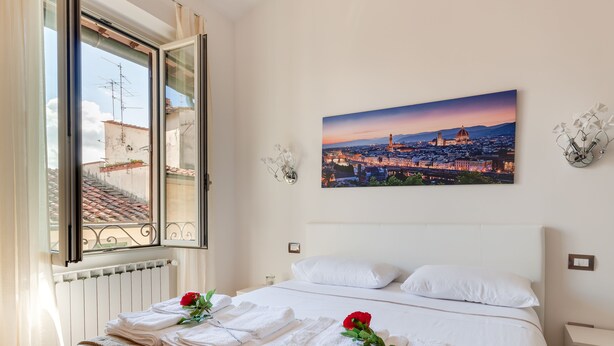 Habitación de hotel con una cama, ropa de cama blanca, una ventana con vista a la ciudad y un cuadro en la pared.