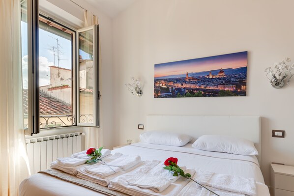 A hotel room with a bed, white linens, a window with a cityscape view, and a framed picture on the wall.