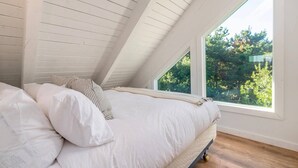 Une chambre avec un plafond en pente, un lit recouvert de draps blancs et une fenêtre donnant sur un paysage verdoyant.