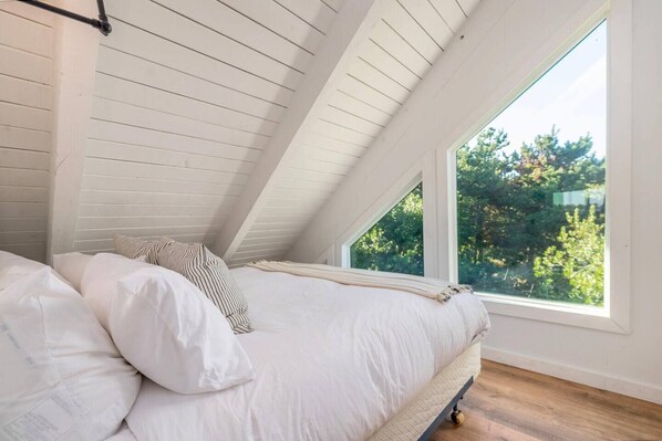 A bedroom with a sloped ceiling, a bed with white bedding, and a window overlooking greenery.