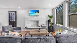 Un salon moderne avec une cheminée en briques blanches, un téléviseur à écran plat fixé au-dessus, et une grande fenêtre offrant une vue sur l’extérieur.
