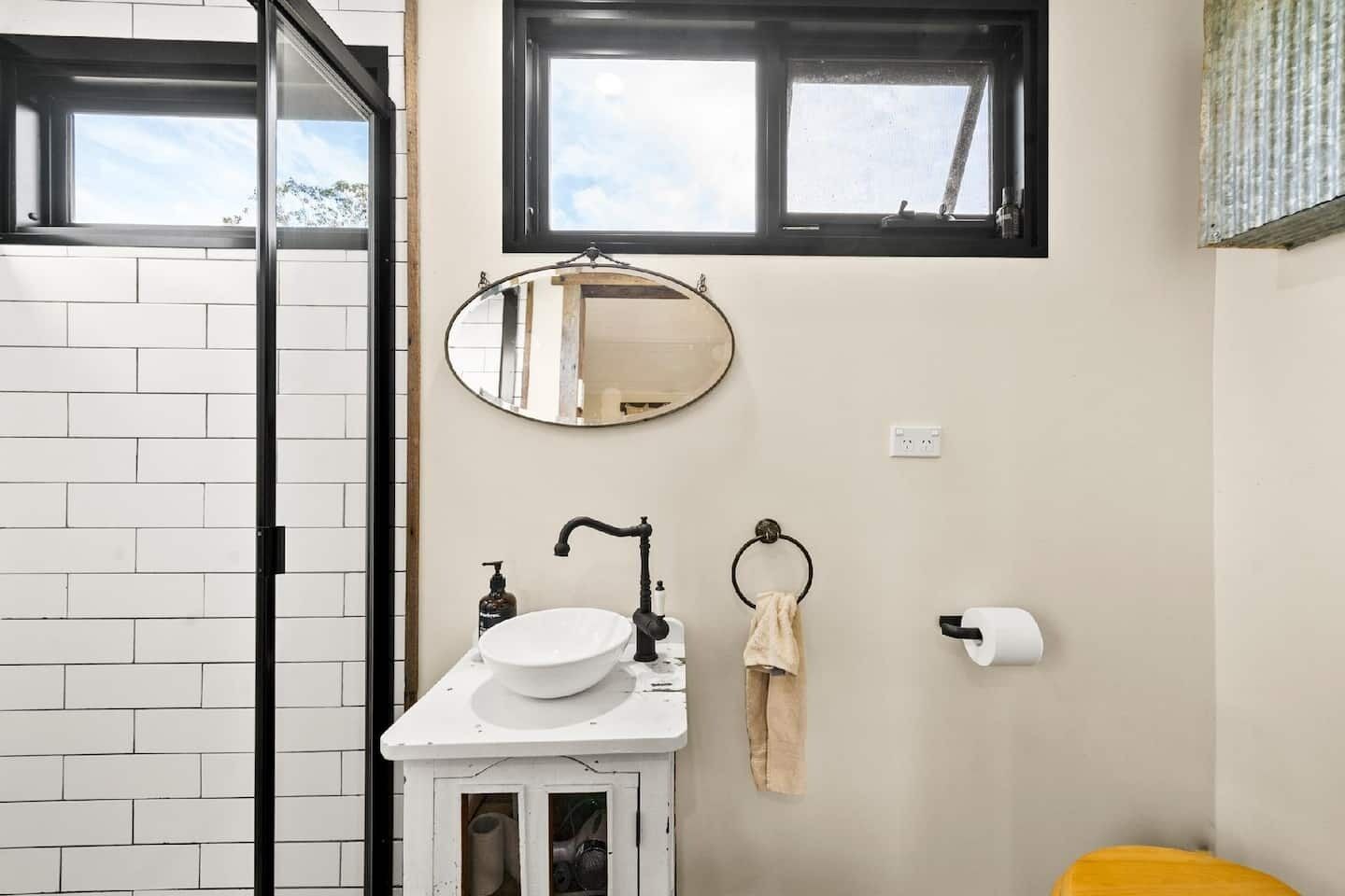 Un baño con un lavamanos blanco, un espejo, un aro para toallas y un dispensador de papel higiénico.