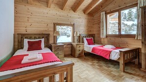 A wooden cabin bedroom with two beds, wooden headboards, and a large window with a view of trees.