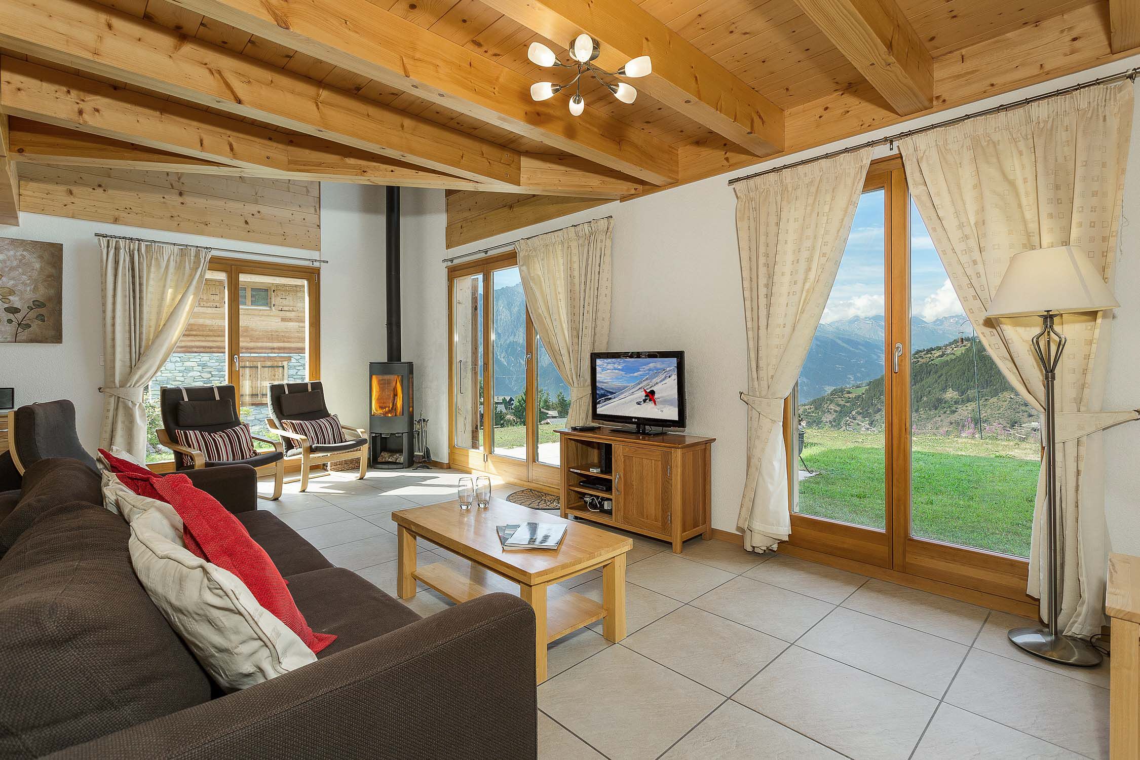 Un salon avec un plafond en bois, un téléviseur, un canapé, une table basse et une vue sur les montagnes, visible à travers de grandes fenêtres.