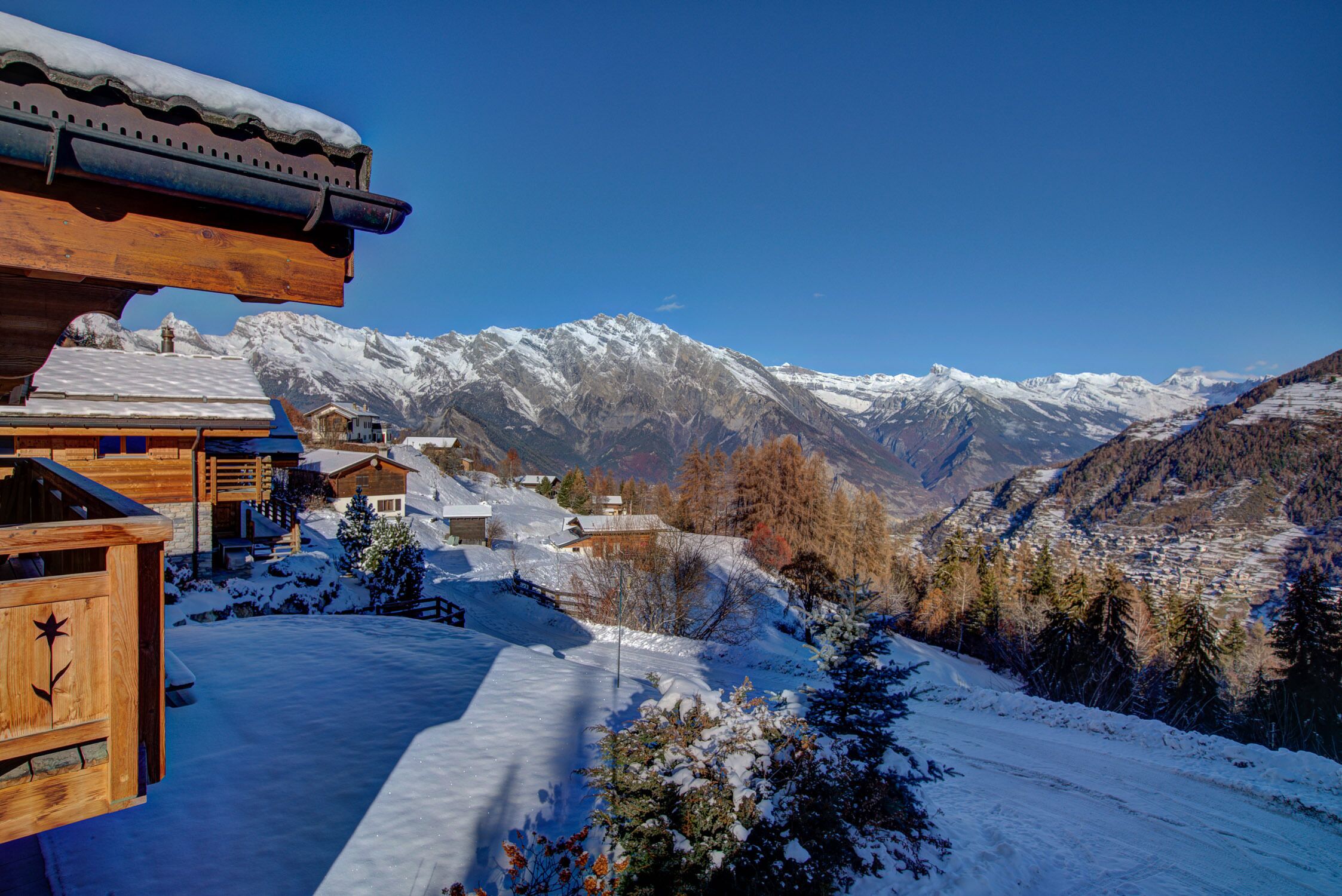 Een houten chalet met een met sneeuw bedekte dak, omringd door bergen en een helderblauwe lucht.