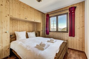 A wooden room with a bed, a window with curtains, and a view of mountains.