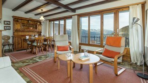Une pièce chaleureuse avec une table à manger en bois, deux chaises, un canapé et de grandes fenêtres donnant sur une vue imprenable sur les montagnes.