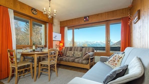Een gezellige kamer met houten inrichting, een zithoek met een tafel en stoelen, een bank en grote ramen met uitzicht op de bergen.