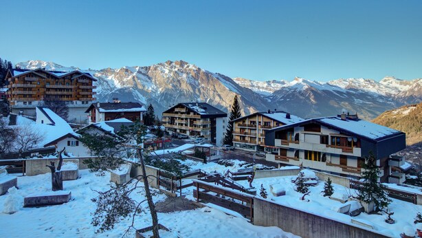 A snow-covered mountain resort with multi-story buildings, ski lifts, and a clear view of snow-capped mountains in the background. - Appartement Bellevue 207 - La Tzoumaz, 4 Vallees (La Tzoumaz)