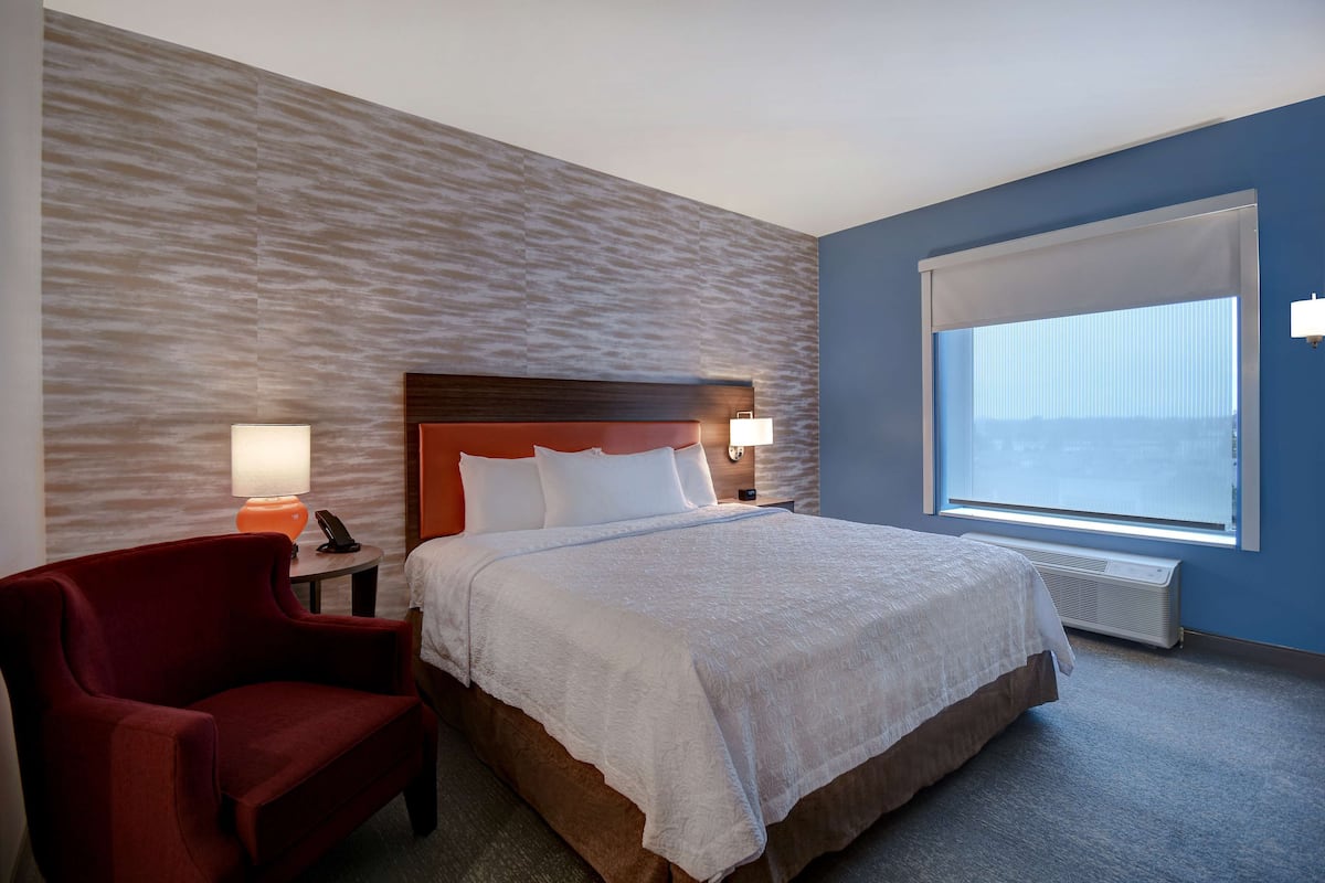 A hotel room with a large bed, a red armchair, a nightstand with a lamp, and a large window with blinds.