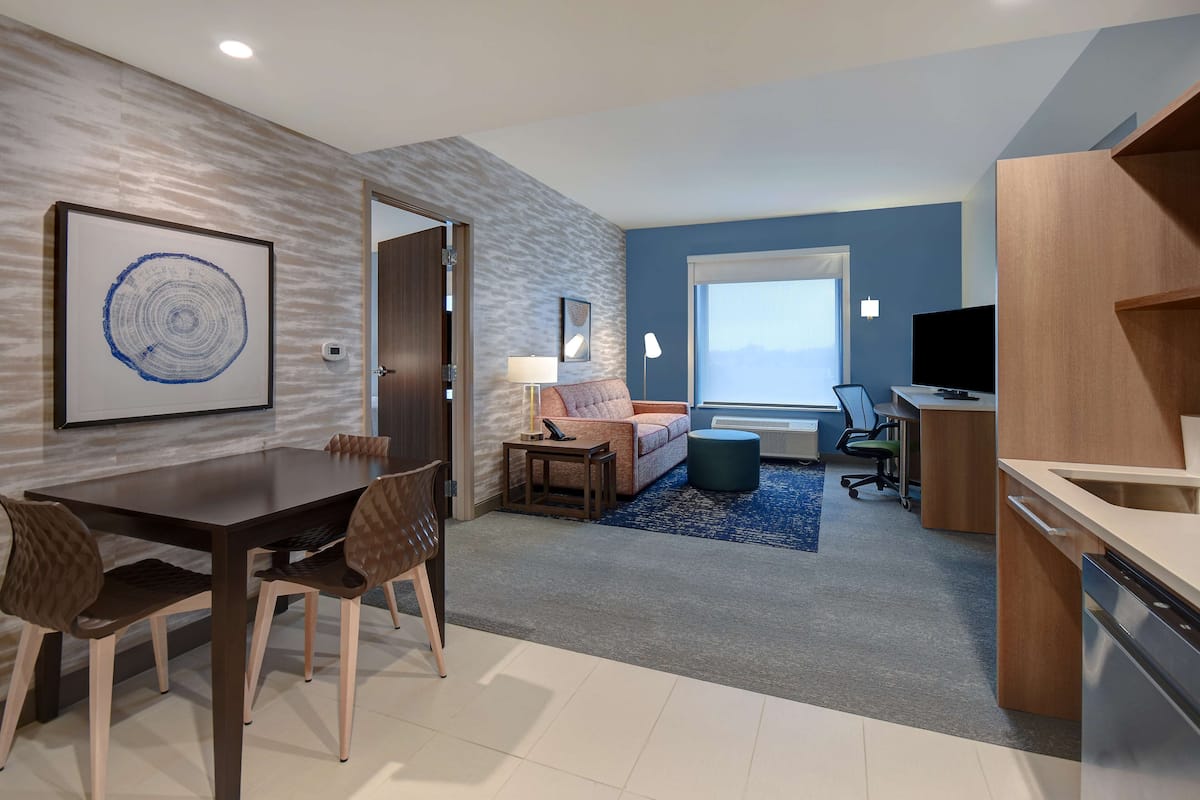 A modern hotel room with a dining area, a sofa, a TV, and a kitchen area.
