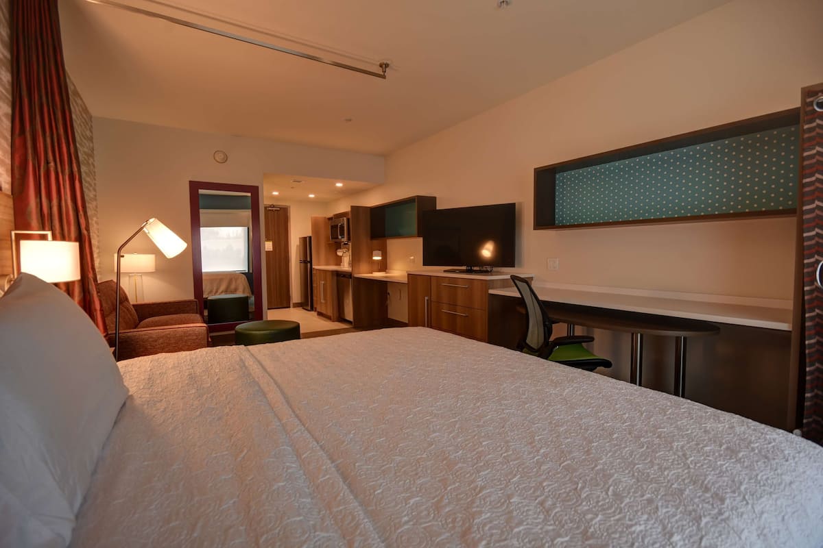A hotel room with a large bed, a desk with a chair, a television, and a small kitchenette.
