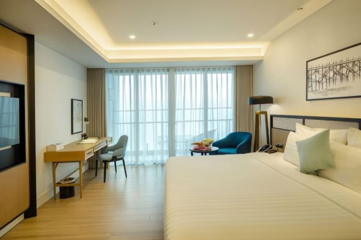 Habitación de hotel con una cama grande, un escritorio, una silla, un televisor y un ventanal con cortinas.