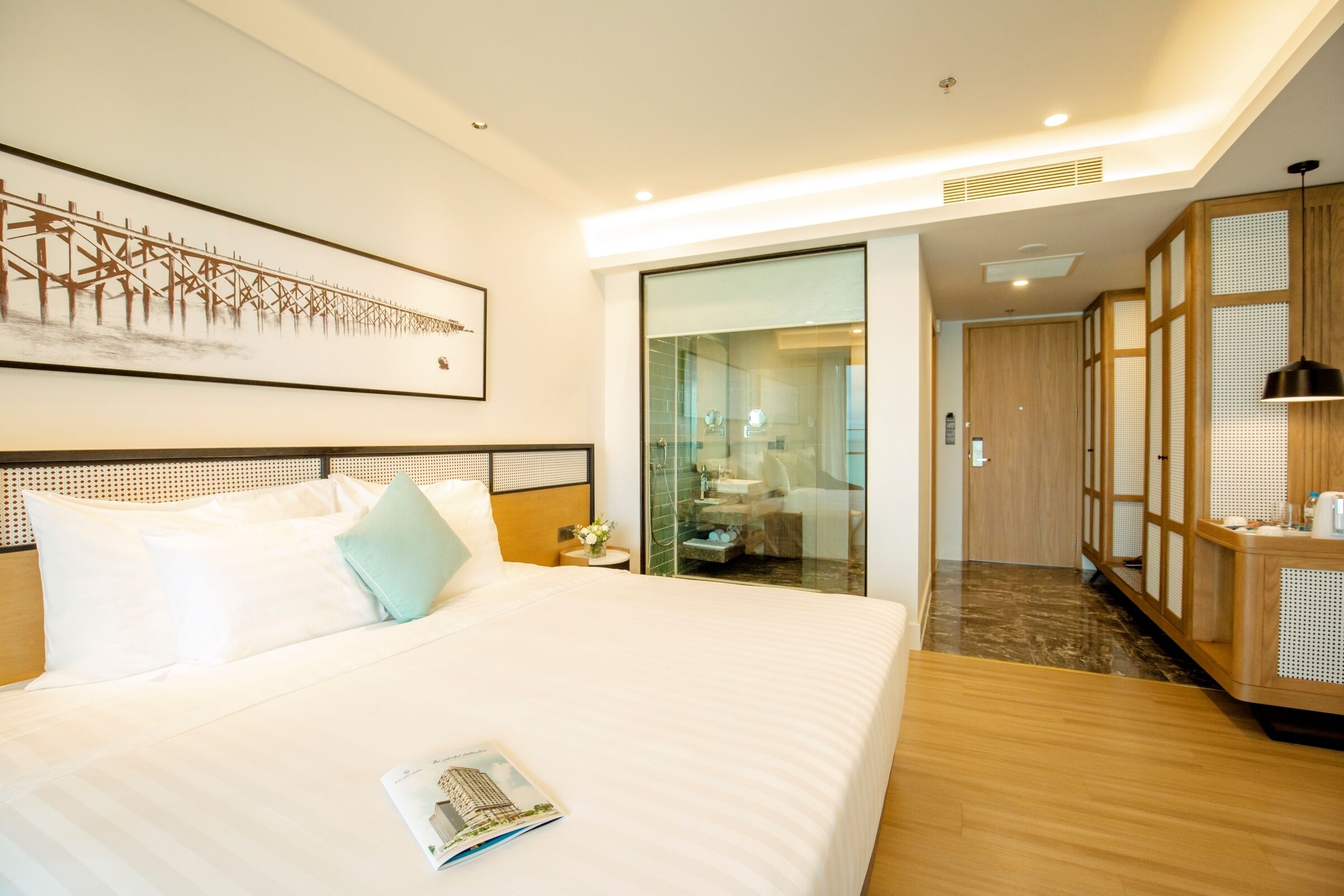 Una habitación de hotel moderna con una cama grande, un separador de vidrio, un armario de madera y una obra de arte enmarcada en la pared.