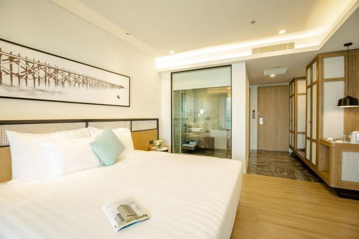 A hotel room with a large bed, a bedside table, a nightstand, a framed artwork on the wall, a sliding glass door leading to a bathroom, and a wooden cabinet.