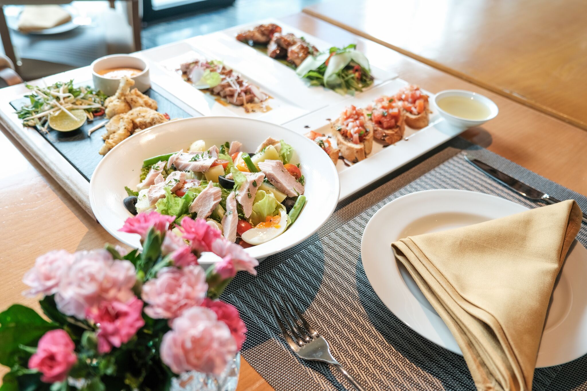 A dining table with various dishes, including a salad, meats, and vegetables, and a vase of pink flowers.