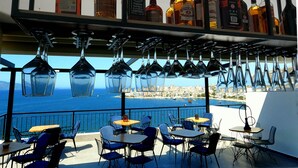 A rooftop bar with a view of the sea and city skyline, featuring multiple tables and chairs, and a variety of liquor bottles on shelves. - Afrodite Suites (Sarandë)