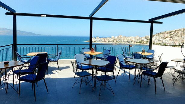 An outdoor terrace with tables and chairs, overlooking a coastal cityscape. - Afrodite Suites (Sarandë)