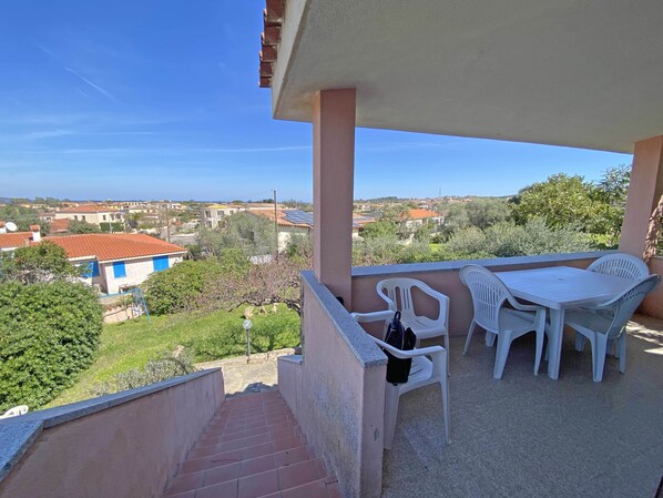 A balcony with a table and chairs, a staircase, and a view of a residential area with houses and greenery.