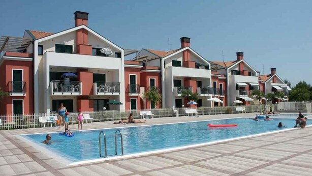 Area piscina con pavimento piastrellato, una vasca con un galleggiante rosso e un edificio con balconi e un recinto bianco.