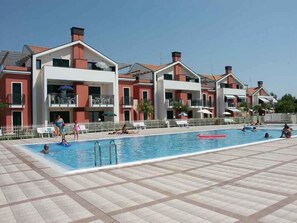 Area piscina con pavimento piastrellato, una vasca con un galleggiante rosso e un edificio con balconi e un recinto bianco.