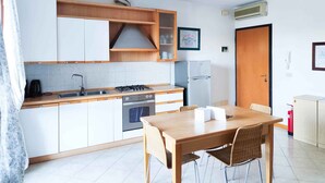 Una cucina compatta con mobili bianchi, piano cottura a gas, forno, lavello e frigorifero. C'è un tavolo da pranzo in legno con sedie.