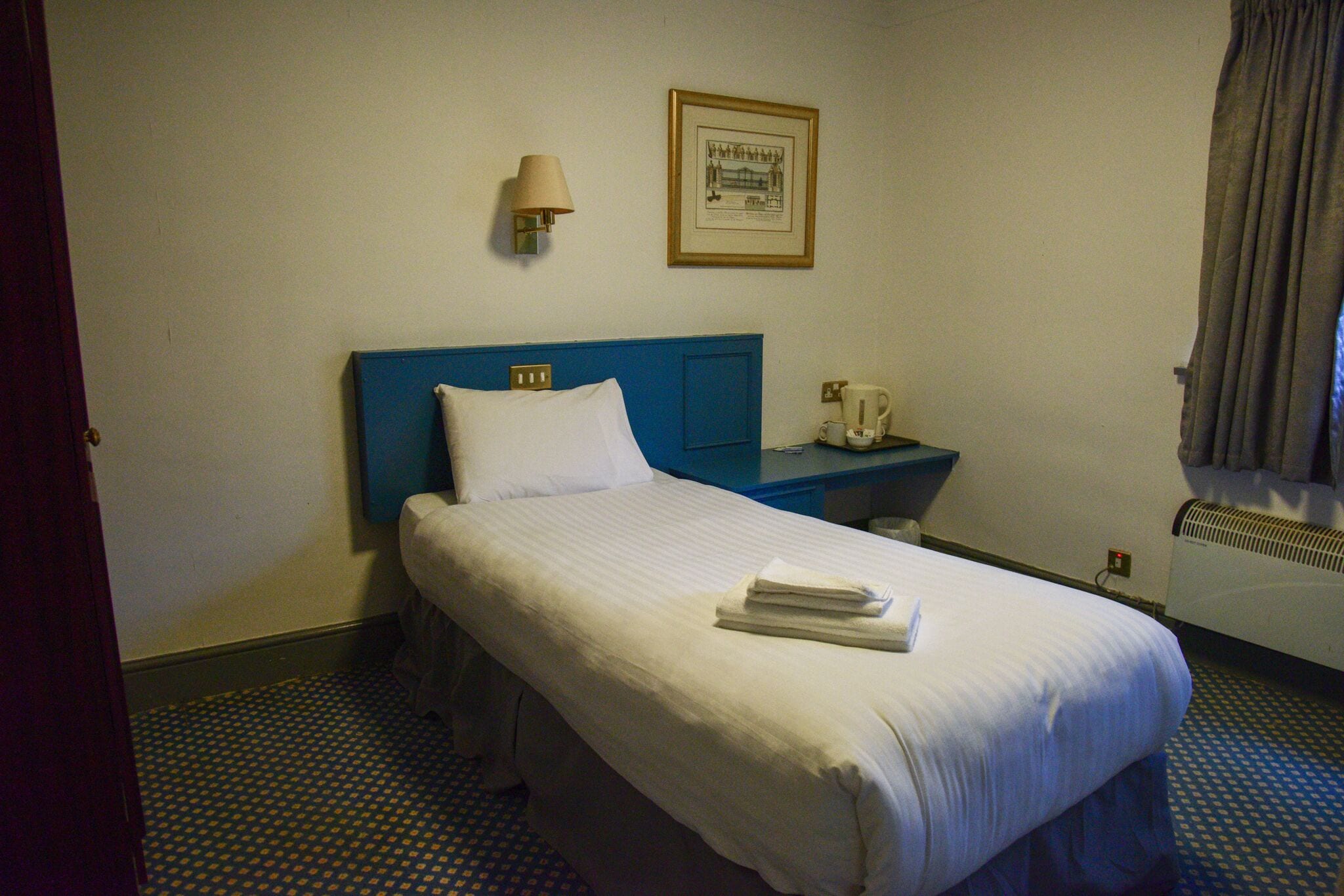 A hotel room with a single bed, a nightstand with a lamp, a framed picture on the wall, and a window with curtains.