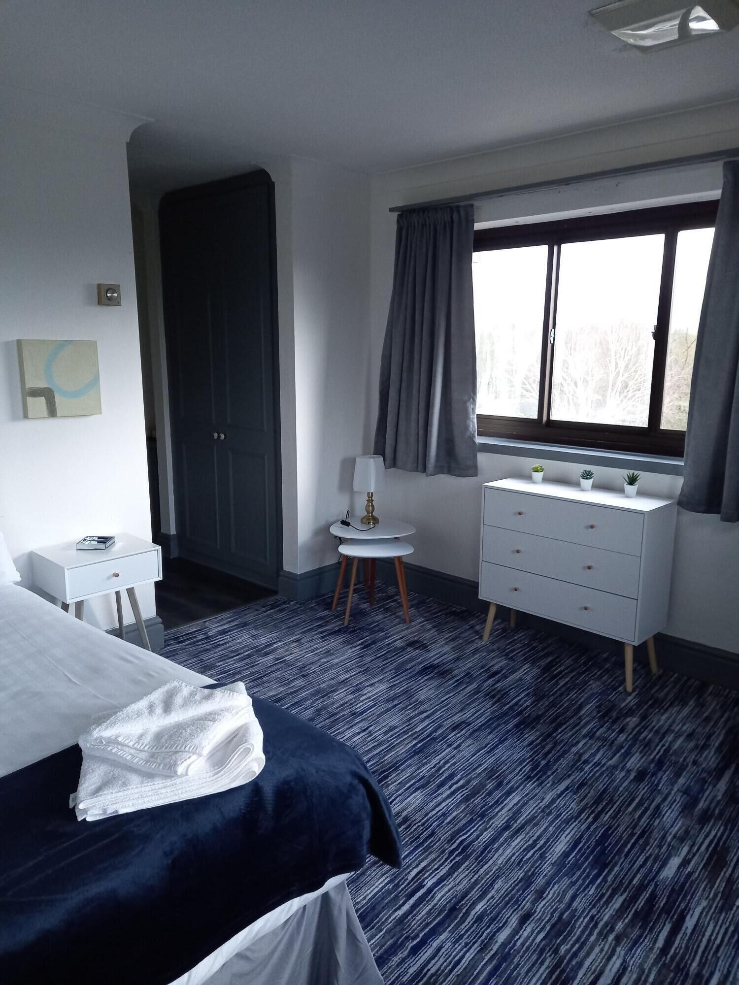 A modern hotel room with a bed, a dresser, a small table with a lamp, and a window with curtains.