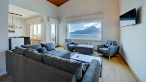A modern living room with a large gray sectional sofa, two armchairs, a coffee table, and a TV mounted on the wall. A large window offers a view of a mountain and the sea. - Casa Bellavista Brenzone (Brenzone)