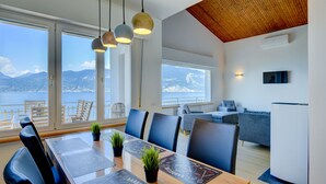 A modern dining area with a wooden table, blue chairs, and potted plants. Large windows offer a view of the sea and mountains. - Casa Bellavista Brenzone (Brenzone)