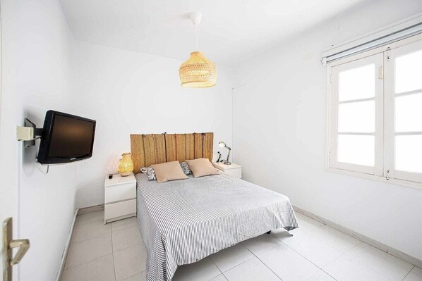 A bedroom with a bed, a television, a bedside lamp, a window, and a hanging light fixture.
