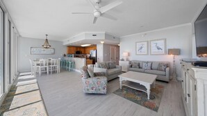 A spacious living room with a white ceiling fan, a dining area with a table and chairs, a kitchen in the background, and a large window providing natural light. - Corner Condo w/ Panoramic Views of the Gulf & Lagoon | Pool & Sauna | Sleeps 10 (Gulf Shores)