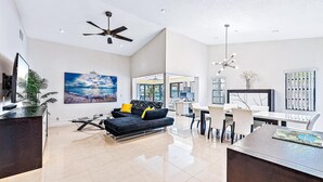 A spacious living area with a modern ceiling fan, a dining table with chairs, a sofa with cushions, and a large window providing natural light.