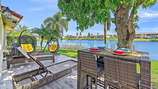 A patio area with wicker furniture, a hanging chair, and a dining table set for a meal by a lake with a building in the background.