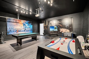 A billiards table, a pool table, and a foosball table in a room with dark walls and a large screen displaying a video game.