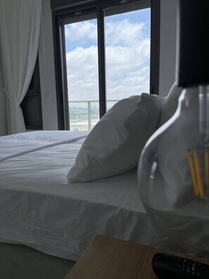 A hotel room with a bed, a nightstand, and a window with a view of the sky and clouds. - LuxuryLiving at Achziv with AmazingViews (Nahariya)