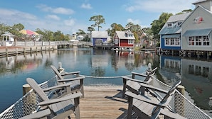 A wooden dock with chairs, a calm water body, and buildings with a rustic architectural style. - Market Street Inn 434 - Studio with 2 queen beds, close to Baytowne Wharf! (Miramar Beach)