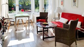 Una habitación luminosa con un sofá rojo, sillas de mimbre, una mesa de comedor y ventanas grandes con cortinas.
